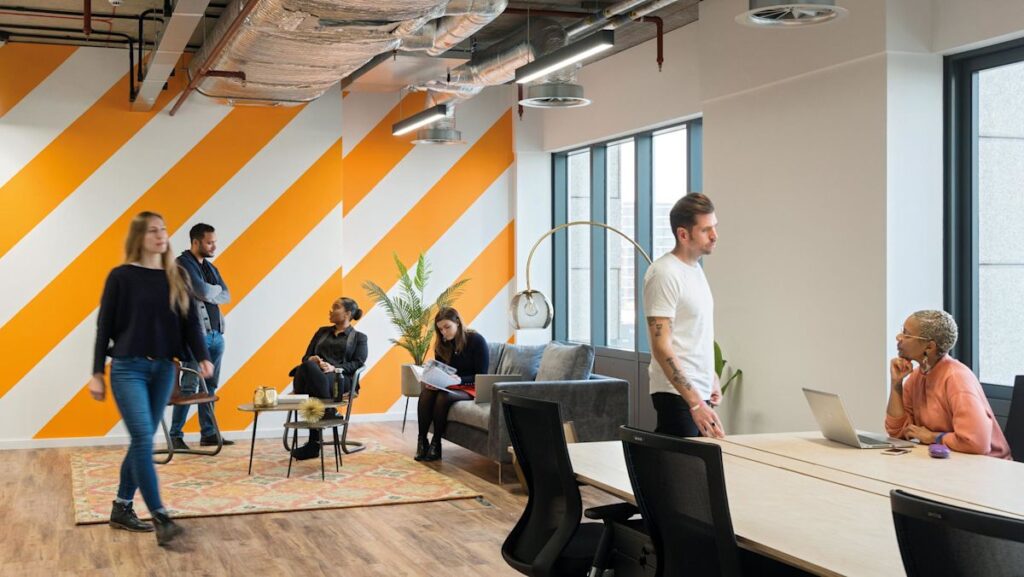 A modern office space with people working and socializing near large windows and an orange-striped wall.