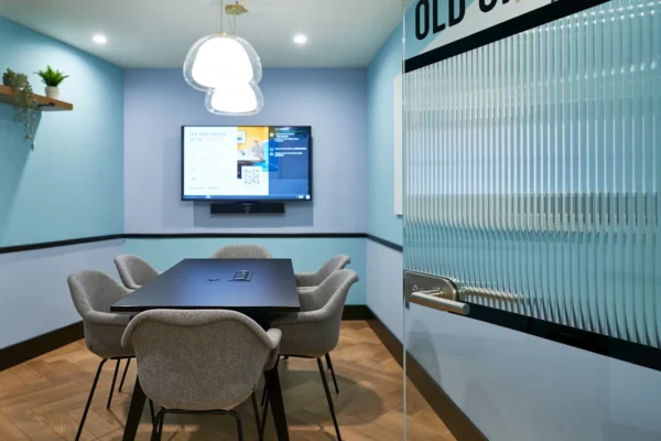 A modern conference room with a rectangular table, four grey chairs, a wall-mounted screen, and a glass door with "OLD" partially visible.