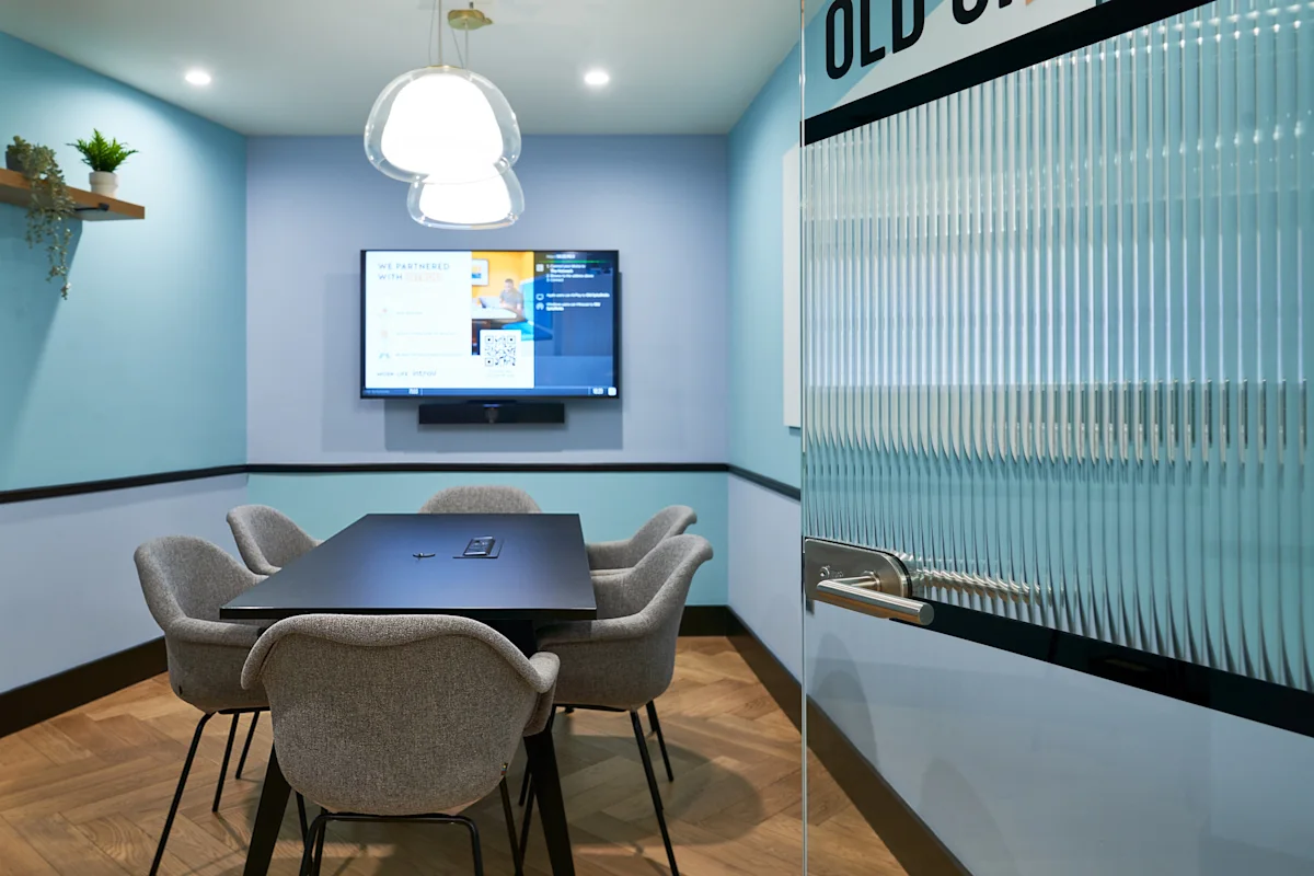 A modern conference room with a rectangular table, four grey chairs, a wall-mounted screen, and a glass door with "OLD" partially visible.