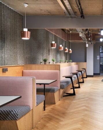 Modern seating area in an office space with wooden tables, pink upholstered booths, and overhead lighting.