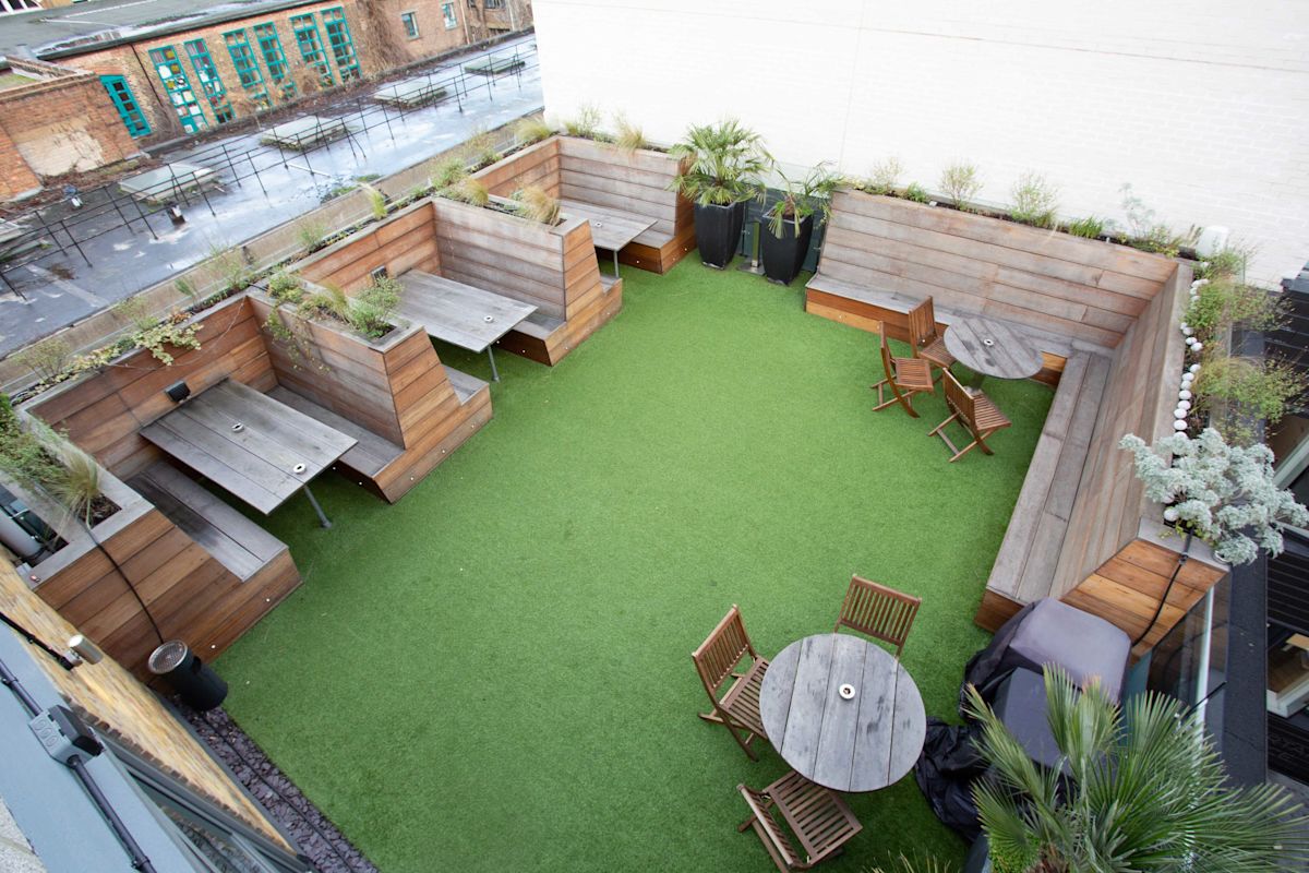 A rooftop terrace with wooden seating areas and tables on green artificial grass at Curtain Road 114 116 in London.