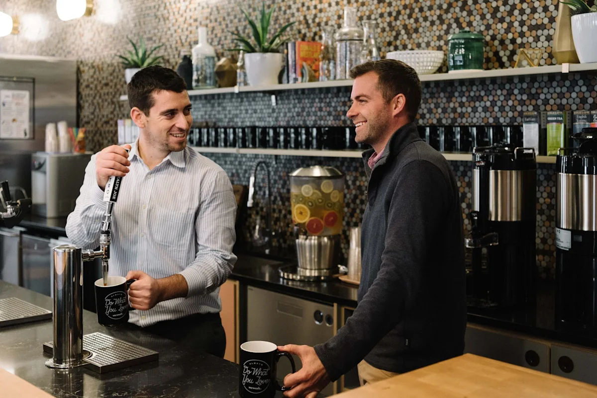 Two men smiling and chatting while preparing drinks at a coffee station in Devonshire Square, London.