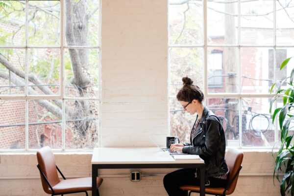 A person works on a laptop at a table by a large window with a view of trees and buildings outside.