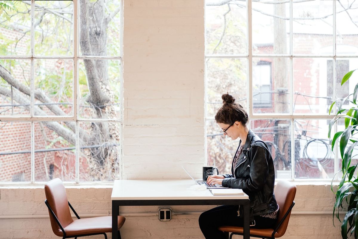 A person works on a laptop at a table by a large window with a view of trees and buildings outside.