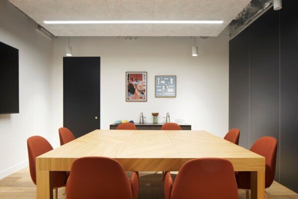 A modern conference room with a large wooden table, orange chairs, artwork on the wall, and minimalist decor.