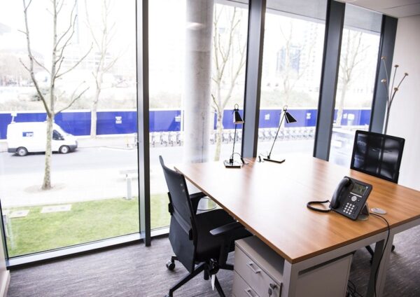 Modern office space with wooden desk, chairs, and large windows overlooking a street in Indescon Square, London.