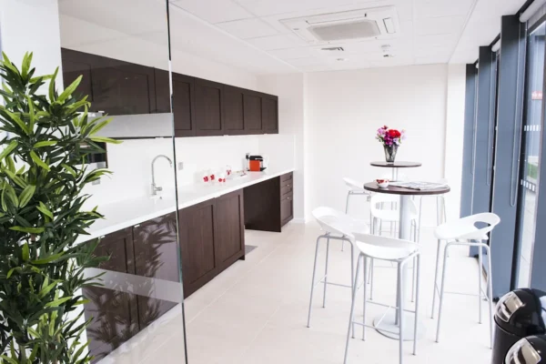 Modern office kitchenette with dark cabinets, high tables, and white chairs at Indescon Square 5 in London.