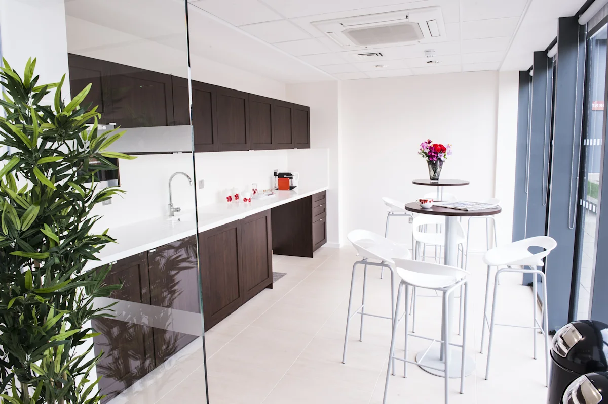 Modern office kitchenette with dark cabinets, high tables, and white chairs at Indescon Square 5 in London.