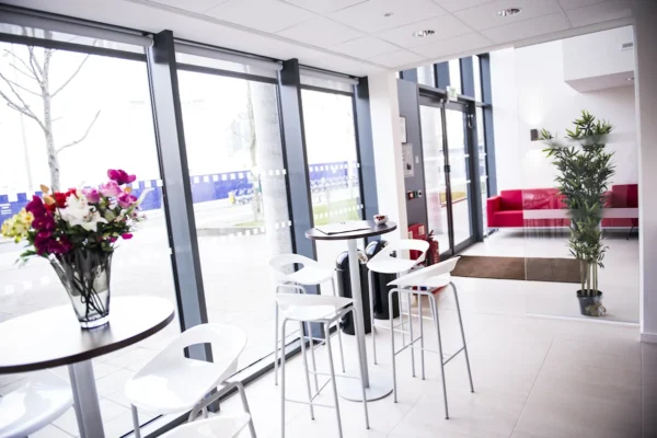A modern lobby area with tall white stools, round tables, and large windows looking outside, featuring a vase of colorful flowers.