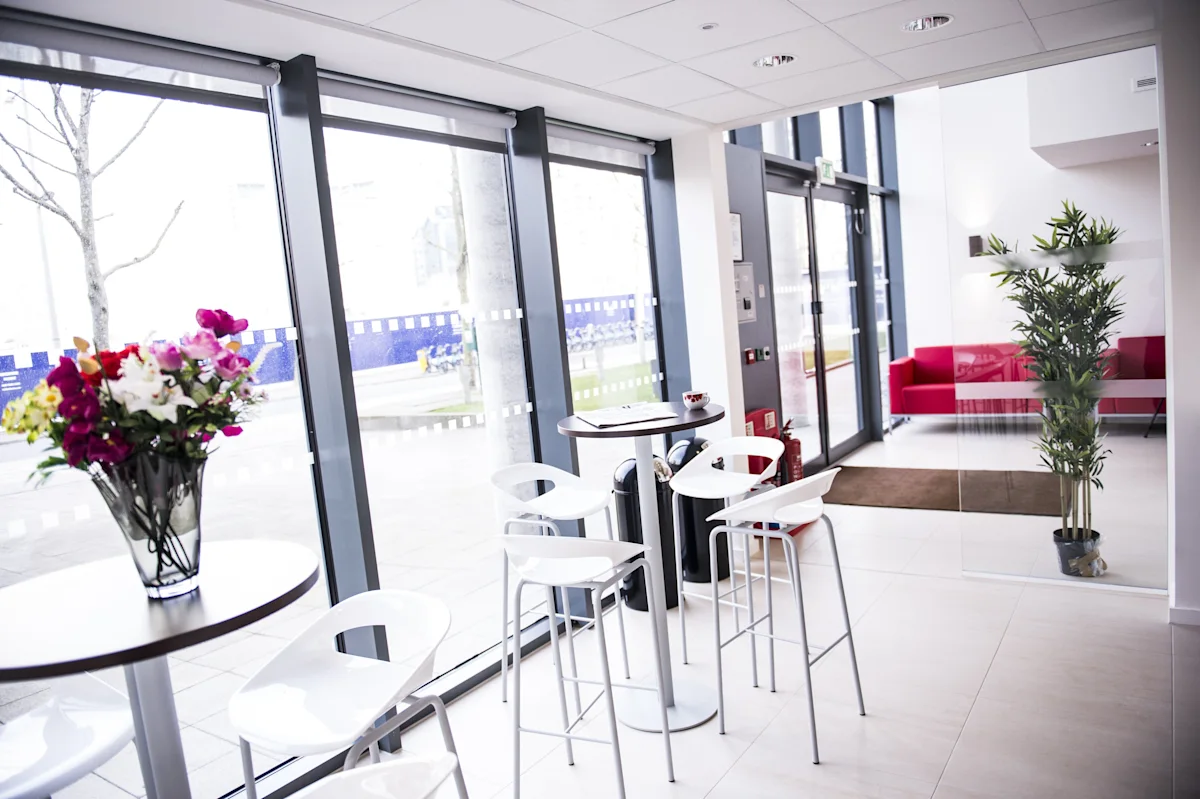 A modern lobby area with tall white stools, round tables, and large windows looking outside, featuring a vase of colorful flowers.
