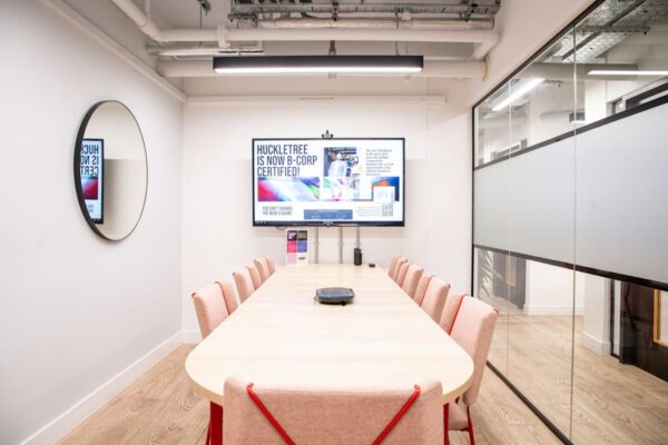 A modern conference room with a wooden table, pink chairs, a large screen displaying "Huckletree is now B-Corp certified," and a circular mirror on the wall.