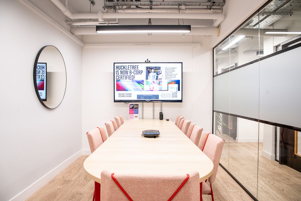 A modern conference room with a wooden table, pink chairs, a large screen displaying "Huckletree is now B-Corp certified," and a circular mirror on the wall.