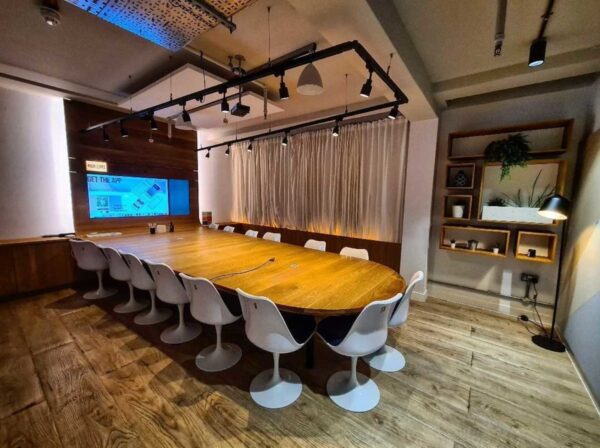 A modern conference room with a large wooden table, white chairs, and a wall-mounted screen.