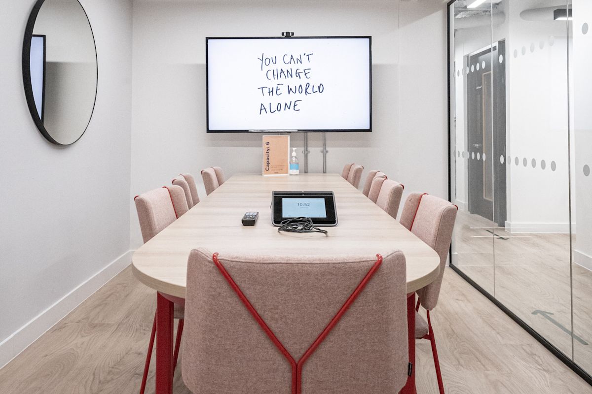 A modern conference room with a large table, a wall-mounted screen displaying "You can't change the world alone," and a mirrored wall.