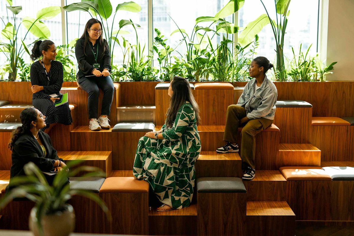 A group of five people sit and talk on wooden tiered seating surrounded by plants in a sunlit room.