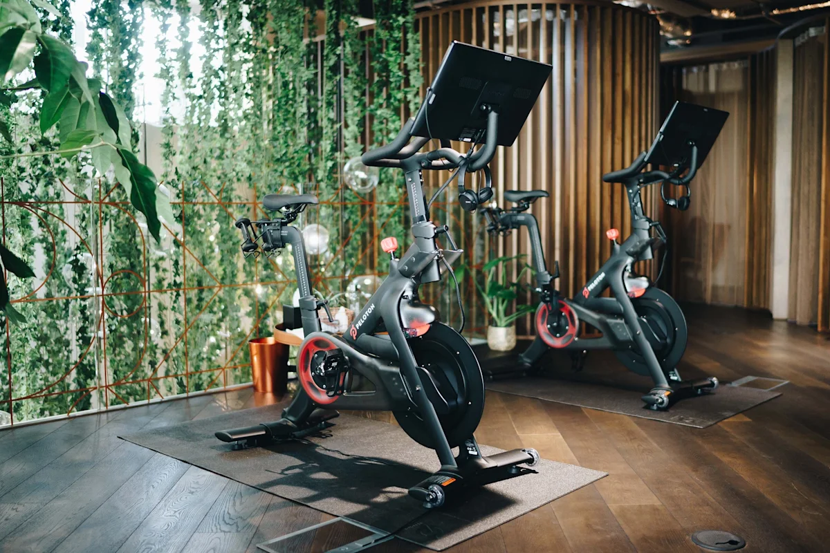 Two Peloton exercise bikes in a stylish gym with wooden flooring and green plant decor.