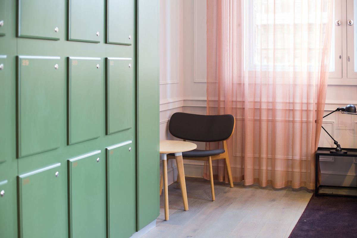 A small office corner with green lockers, a black chair, a light wooden table, and pink curtains.