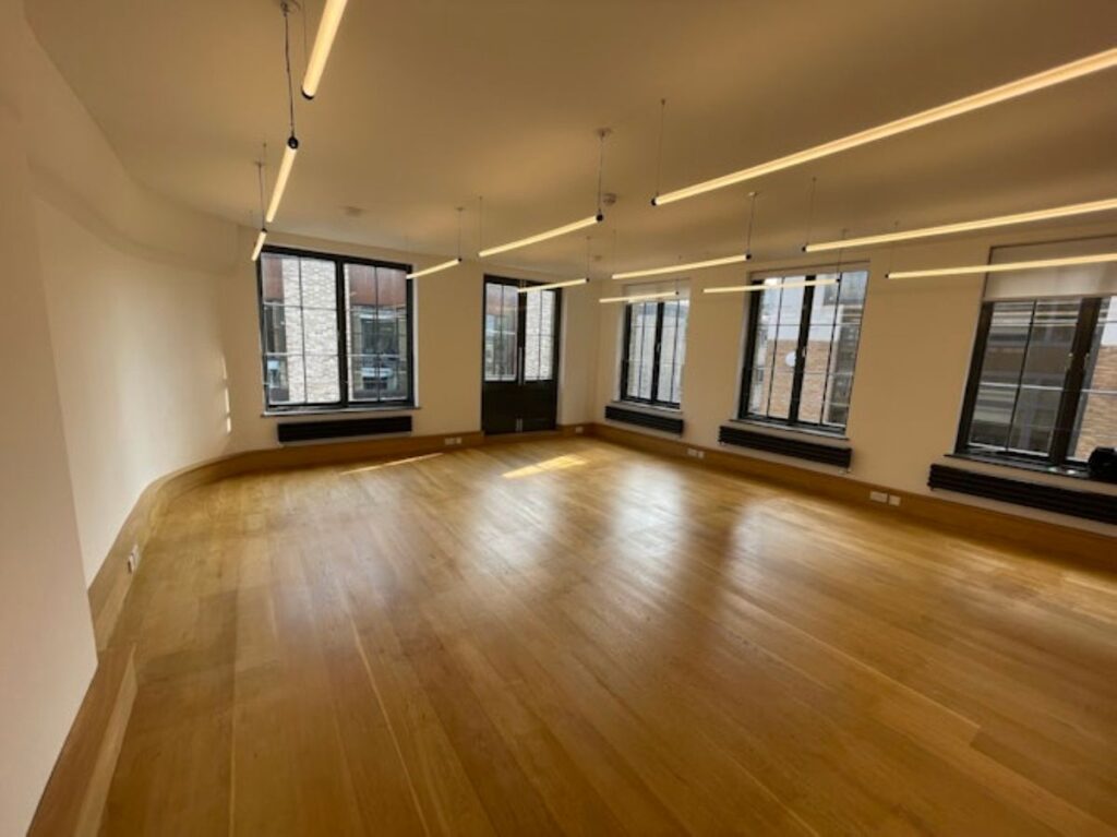 An empty office space with wooden floors, large windows, and ceiling lights at Luke Street 78a, London.