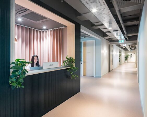 A modern office reception area at Marsh Wall 77 London, featuring a woman working at a desk with plants and hallway in the background.