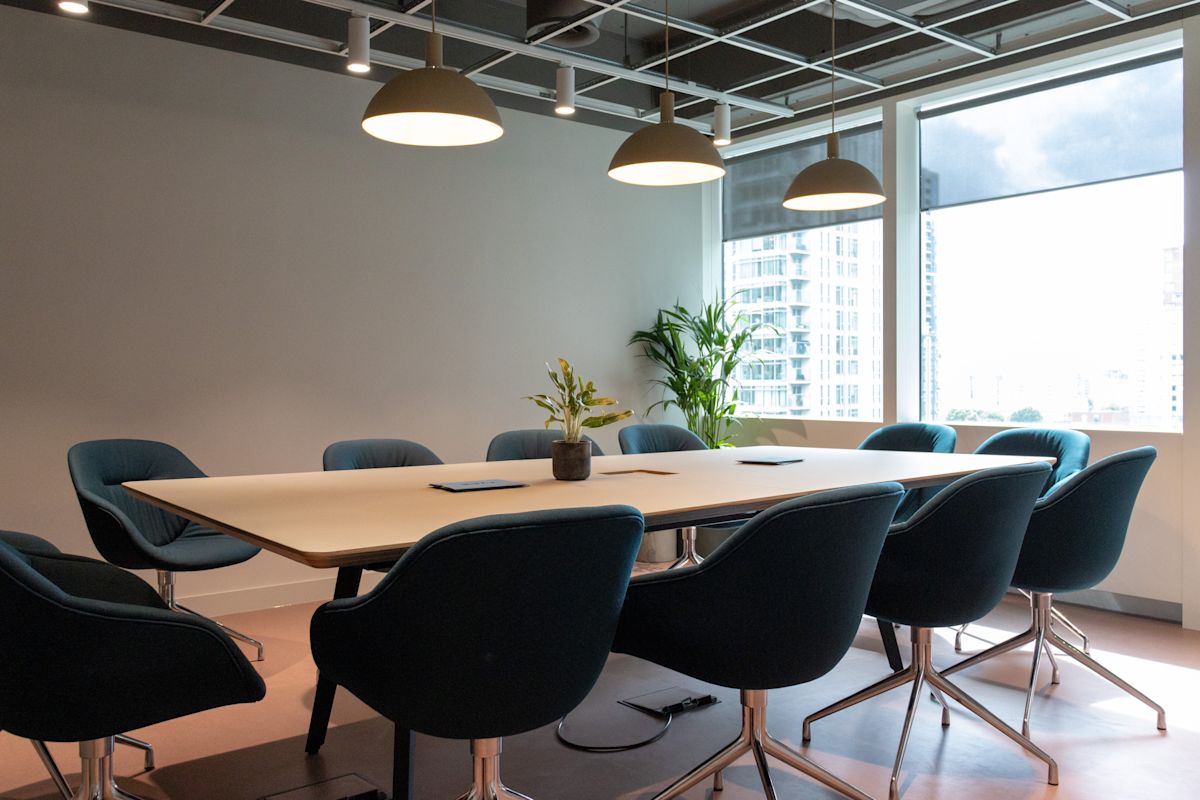 A modern conference room with a large table, surrounded by dark chairs, illuminated by ceiling lights, with a view of city buildings from the window.