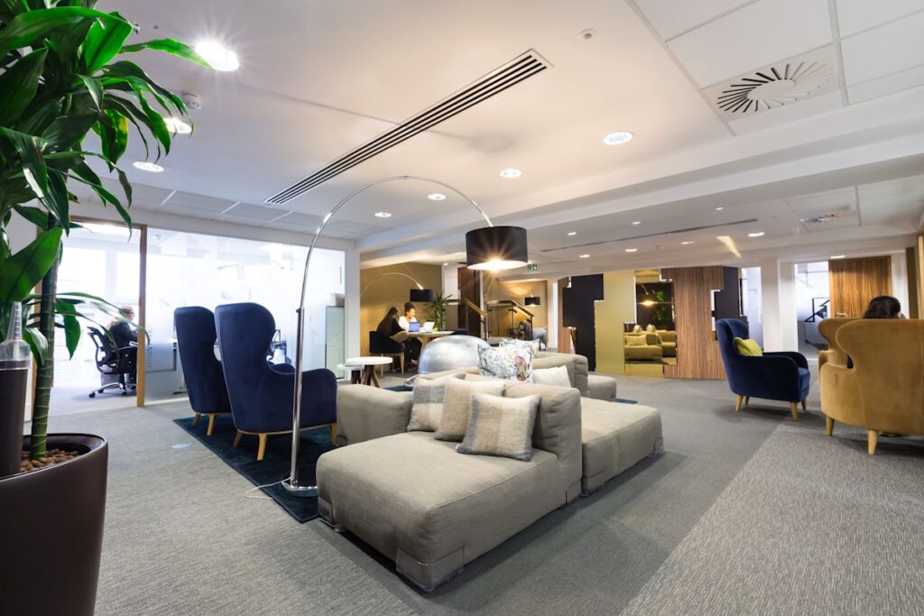 A modern office lounge area with comfortable seating, plants, and people working on laptops.
