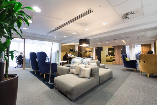 A modern office lounge area with comfortable seating, plants, and people working on laptops.