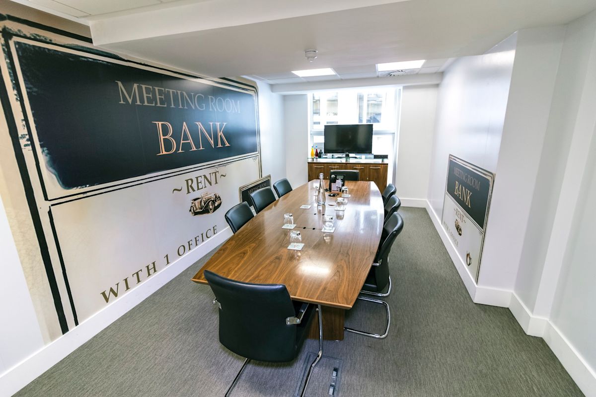 A modern meeting room with a long wooden table, surrounded by chairs and featuring a sign reading "Meeting Room Bank."