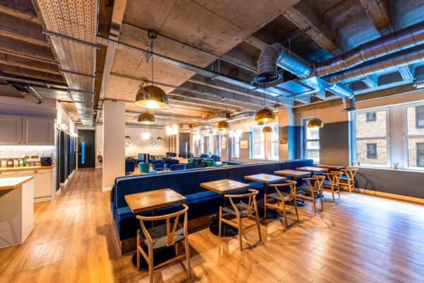 A spacious, modern interior of an office lounge with wooden floors, blue seating, and industrial-style lighting.