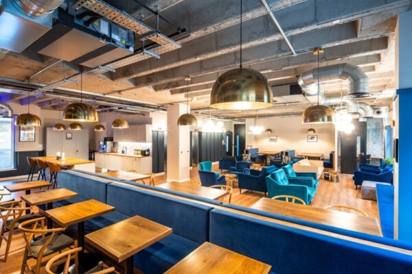 A modern, stylish lounge area with wooden tables, blue seating, and gold dome lighting fixtures in a co-working space.