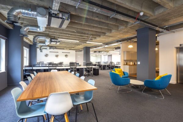 A modern, open-plan office space with a wooden conference table, colorful chairs, and industrial ceiling design.