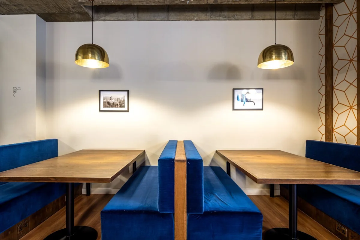 A modern seating area with blue velvet benches and wooden tables under brass pendant lights.