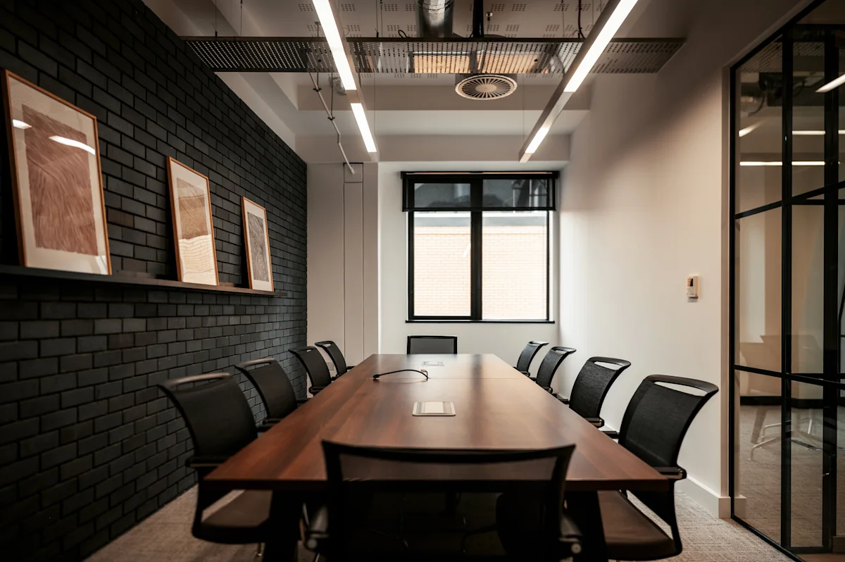 A modern conference room with a long wooden table, black chairs, and framed artwork on a brick wall.