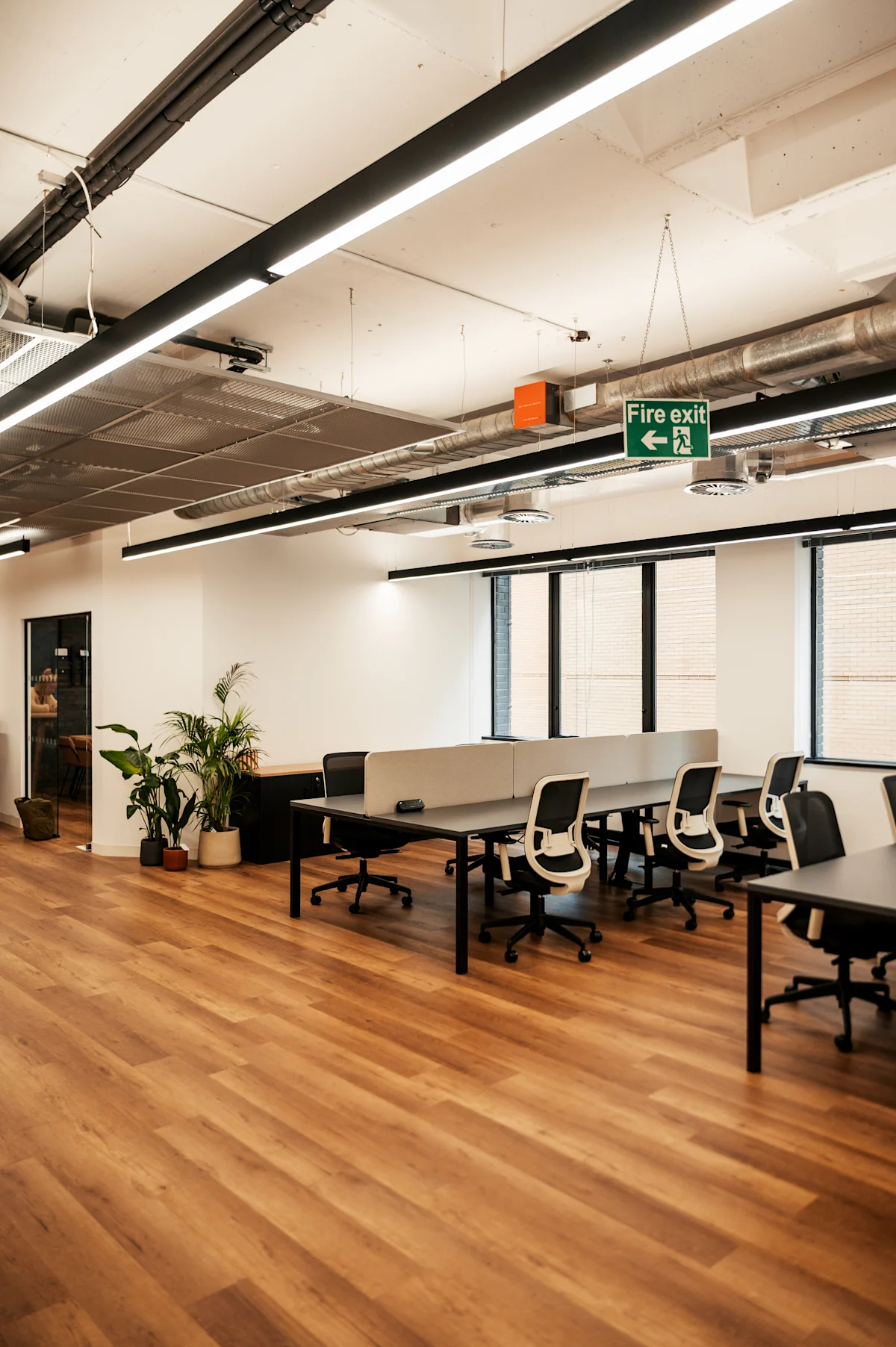 Modern open-plan office space with desks, chairs, plants, and a fire exit sign, located in Norton Folgate, London.