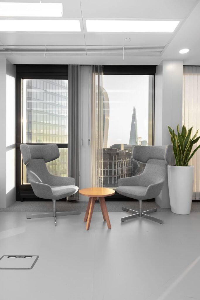 Two modern grey chairs and a small wooden table in an office space with a view of London skyscrapers in the background.