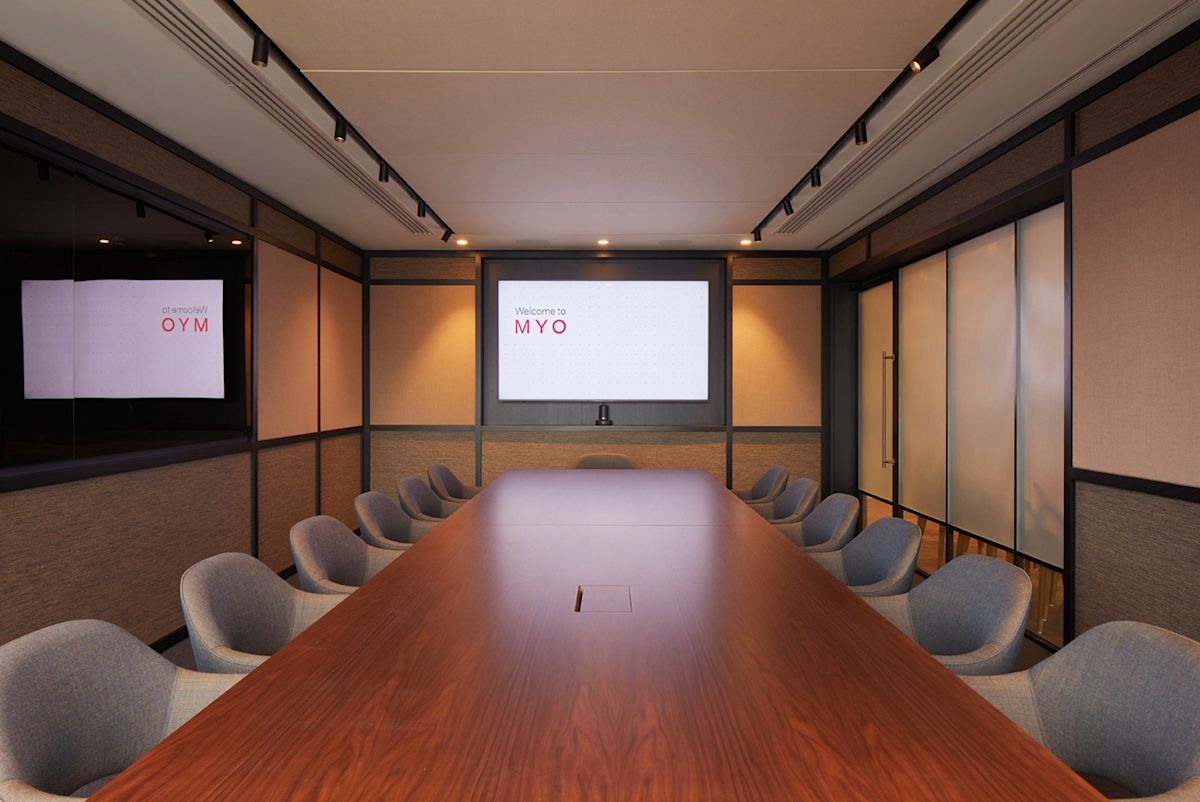 A modern conference room with a large wooden table and a screen displaying "Welcome to MYO."