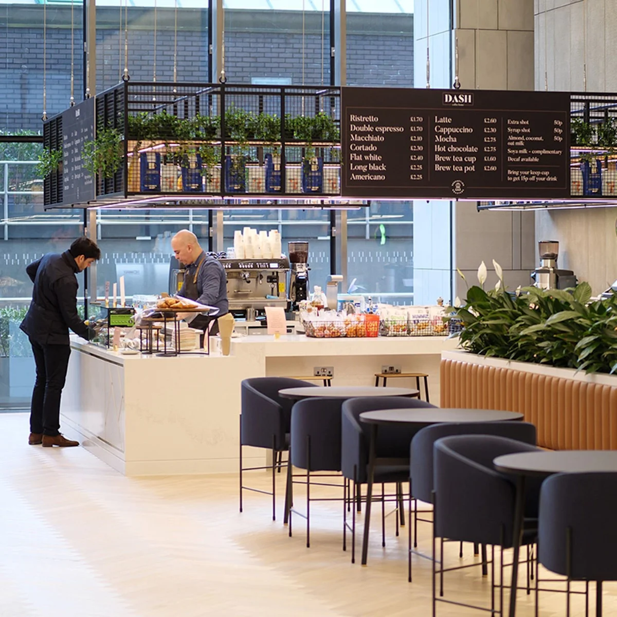 A modern cafe interior on Old Broad Street, London, featuring a central counter with a barista and a customer.