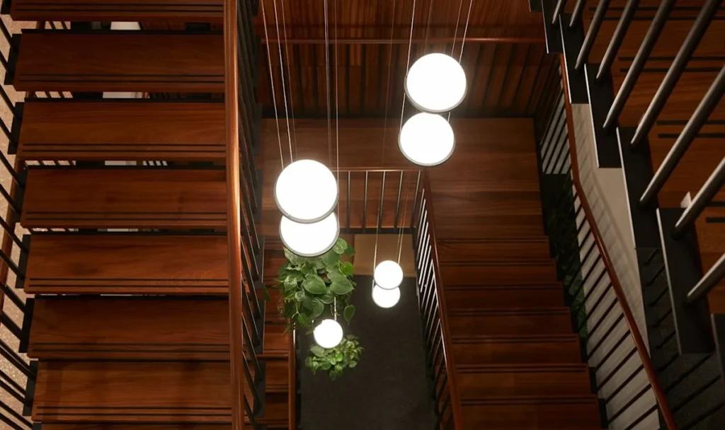 A view down a wooden spiral staircase illuminated by globe pendant lights, with a hanging plant in the center.