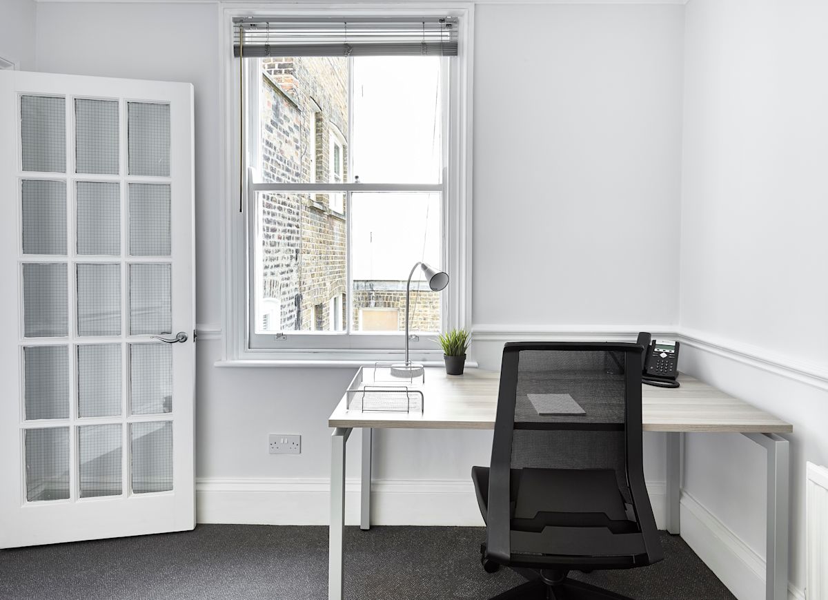 A modern, minimalist office space at Poland Street 54, London featuring a desk with a lamp and phone near a window.