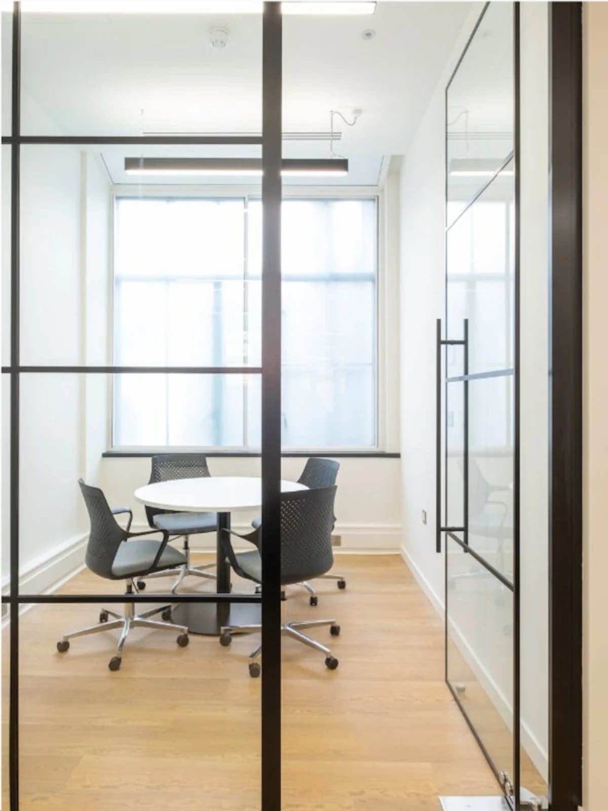 A small, modern conference room with a round table and four chairs seen through a glass door.