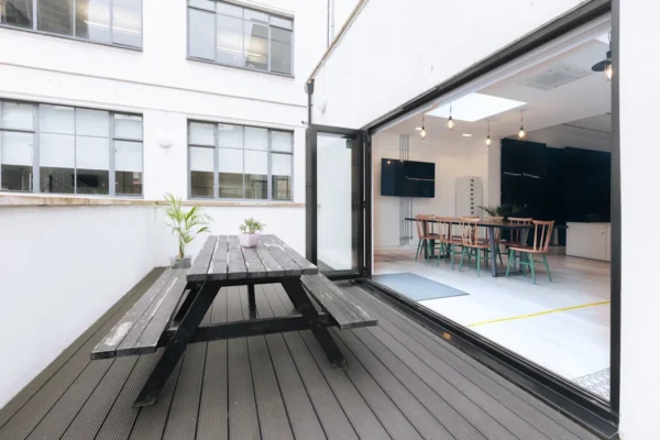 Outdoor terrace with a wooden picnic table and view into a modern indoor meeting space through large open doors.
