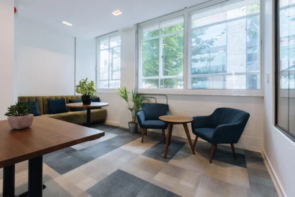 A modern, light-filled office lounge with plants, blue armchairs, and a wooden table.