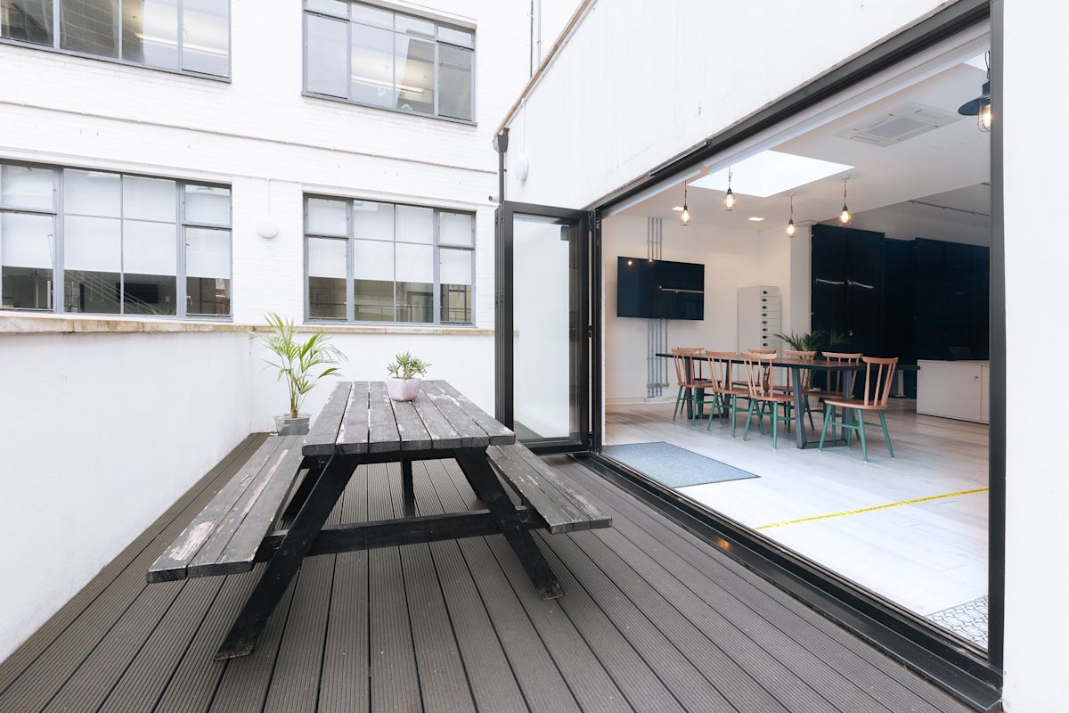 Outdoor terrace at Scrutton Street 32-38, London, featuring a wooden picnic table and a view into a modern office interior.