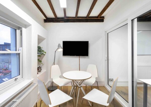 A small, modern meeting room with a round table, white chairs, and a wall-mounted TV.