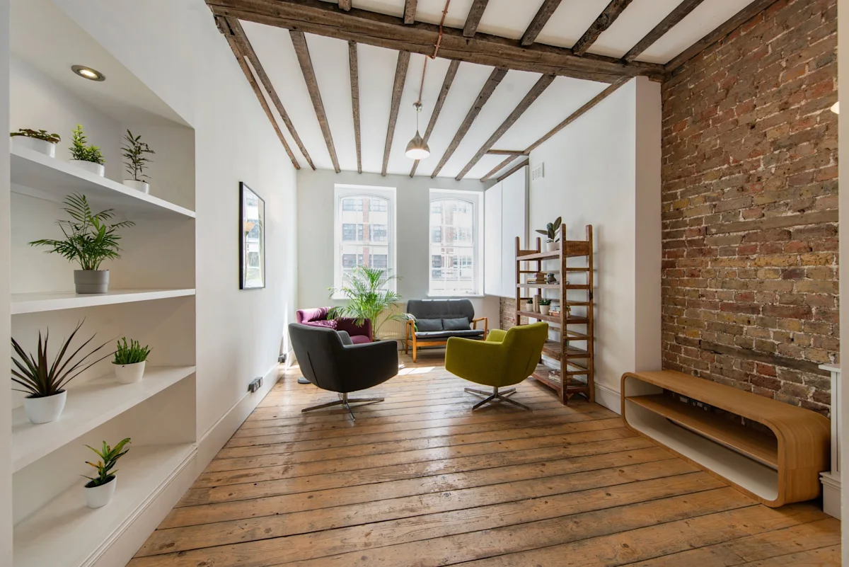 A cozy living room with wooden floors, modern chairs, shelves with plants, and large windows.