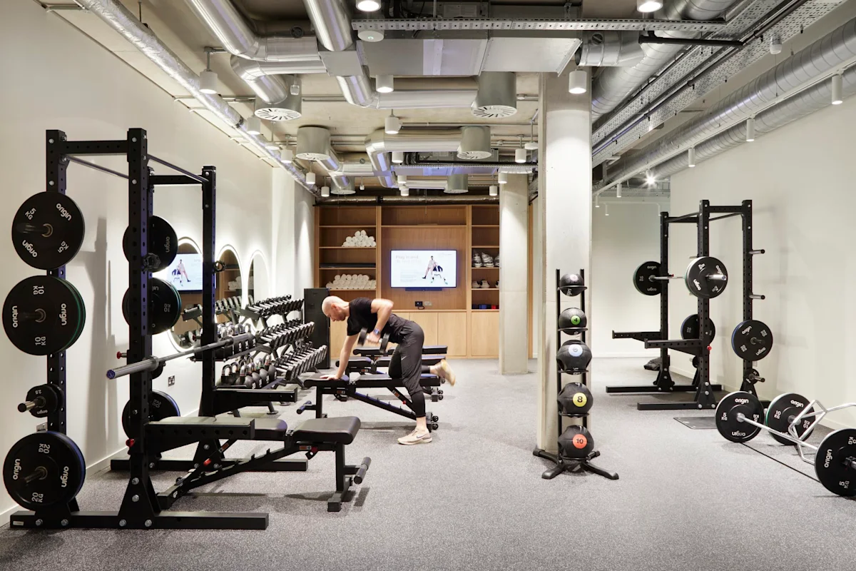 A modern gym in Shoreditch High Street, London, featuring weightlifting equipment and a person exercising.