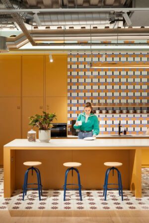 A person in a green sweater stands in a modern kitchen with a yellow and white color scheme at Shoreditch High Street, Montacute Yards, London.
