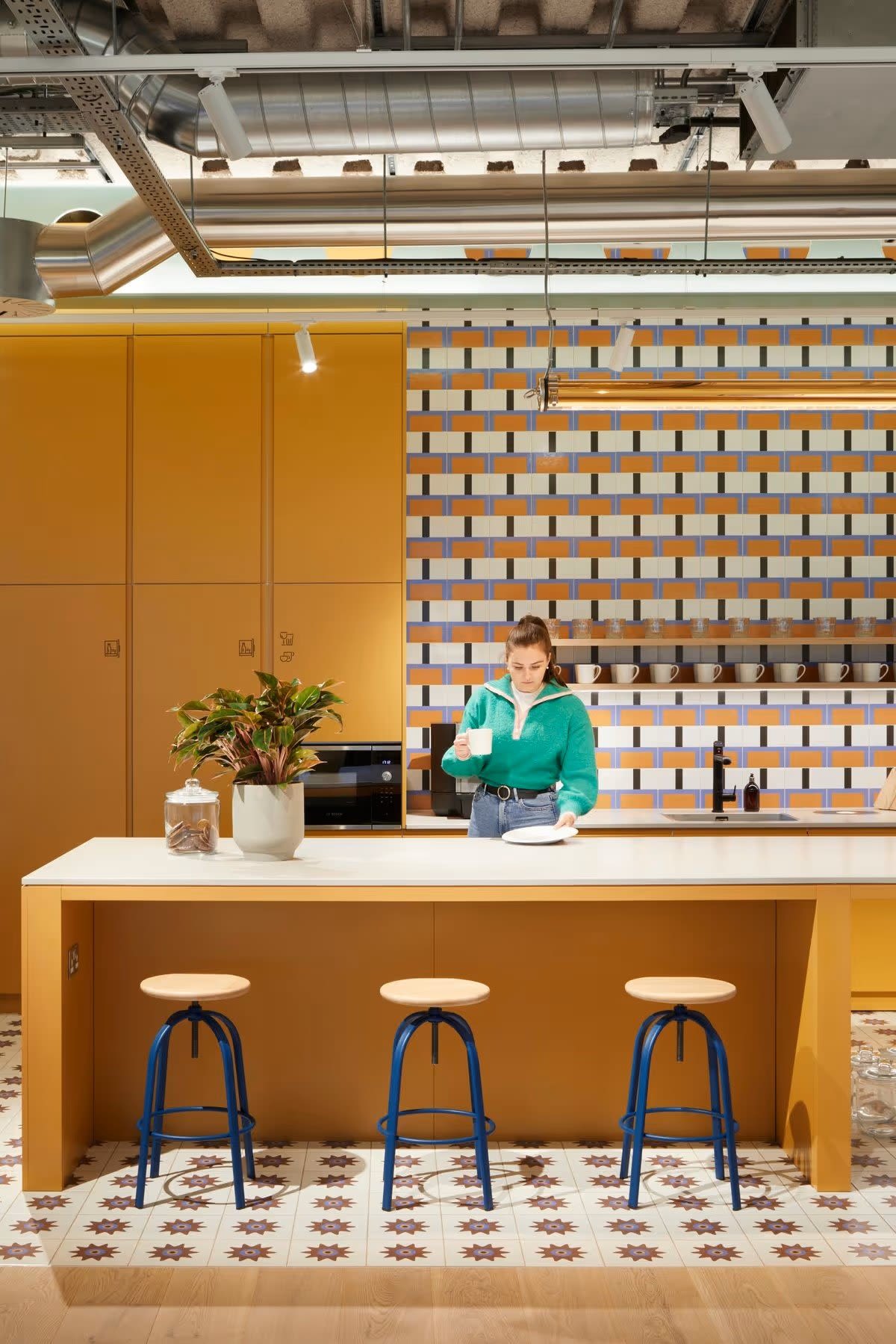 A person in a green sweater stands in a modern kitchen with a yellow and white color scheme at Shoreditch High Street, Montacute Yards, London.