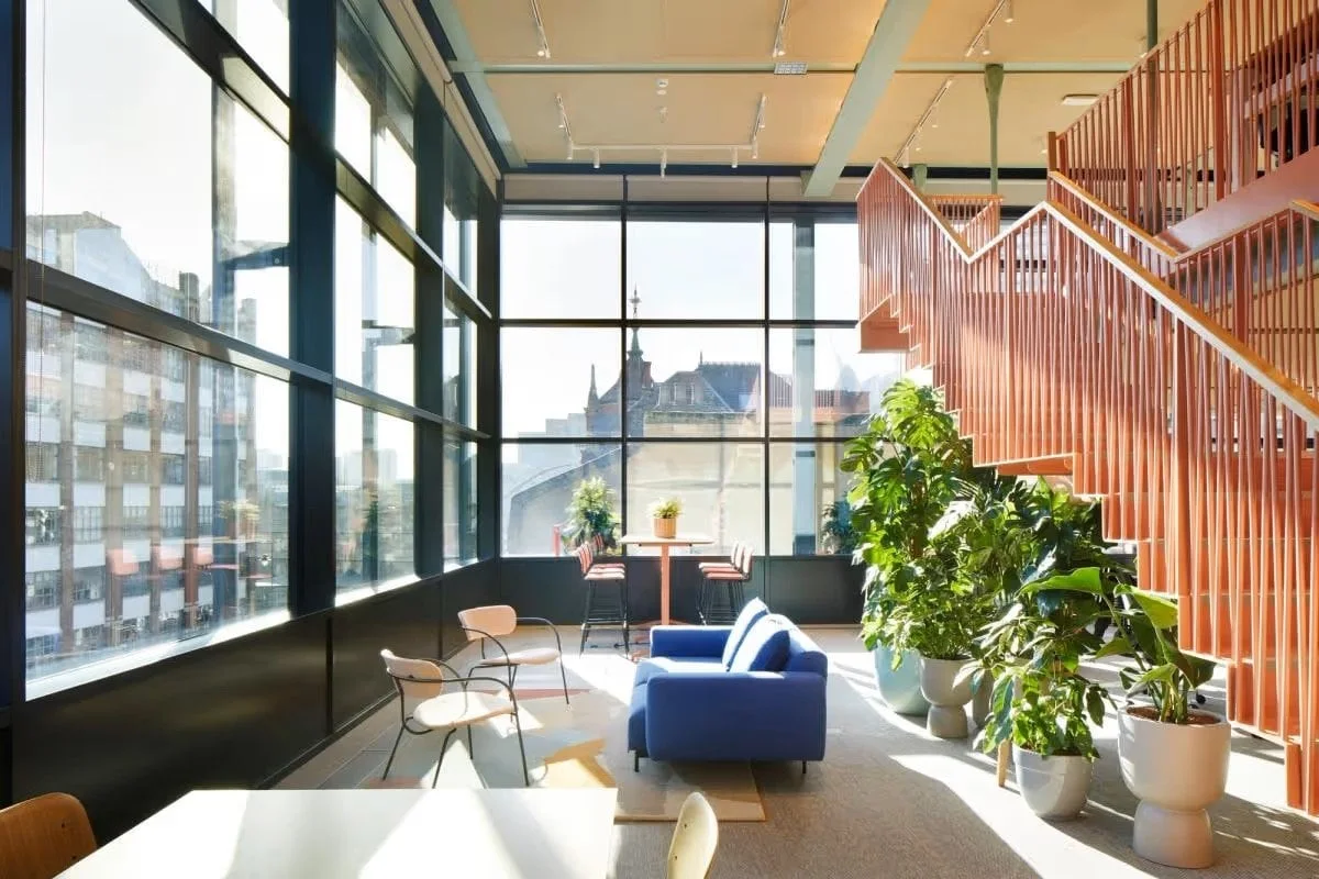 A modern, sunlit office space with large windows, plants, and colorful furniture in Shoreditch High Street, London.