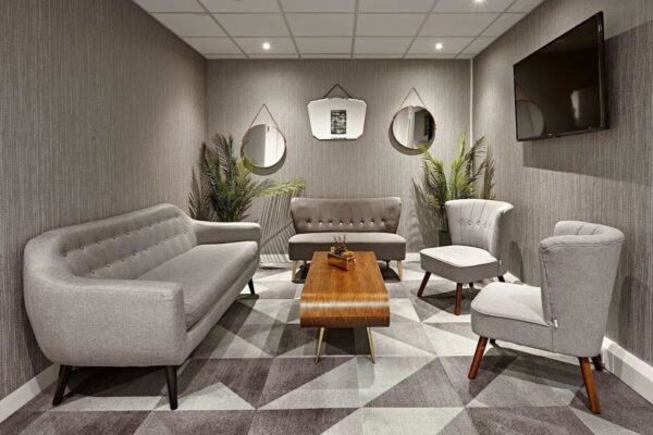 A modern lounge area with grey sofas, armchairs, a wooden coffee table, mirrors, and plants.