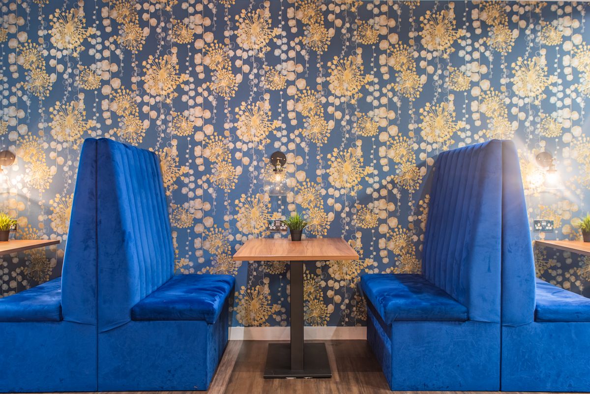 Two blue upholstered booths with a wooden table between them, set against a floral-patterned wallpaper.